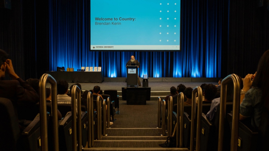 A presenter stands at a lectern delivering a Welcome to Country presentation to a seated audience in a theatre