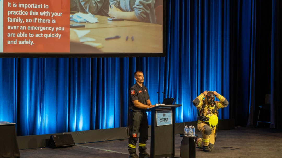 Fire and rescue presenters demonstrate fire safety procedures on stage in front of a large audience.