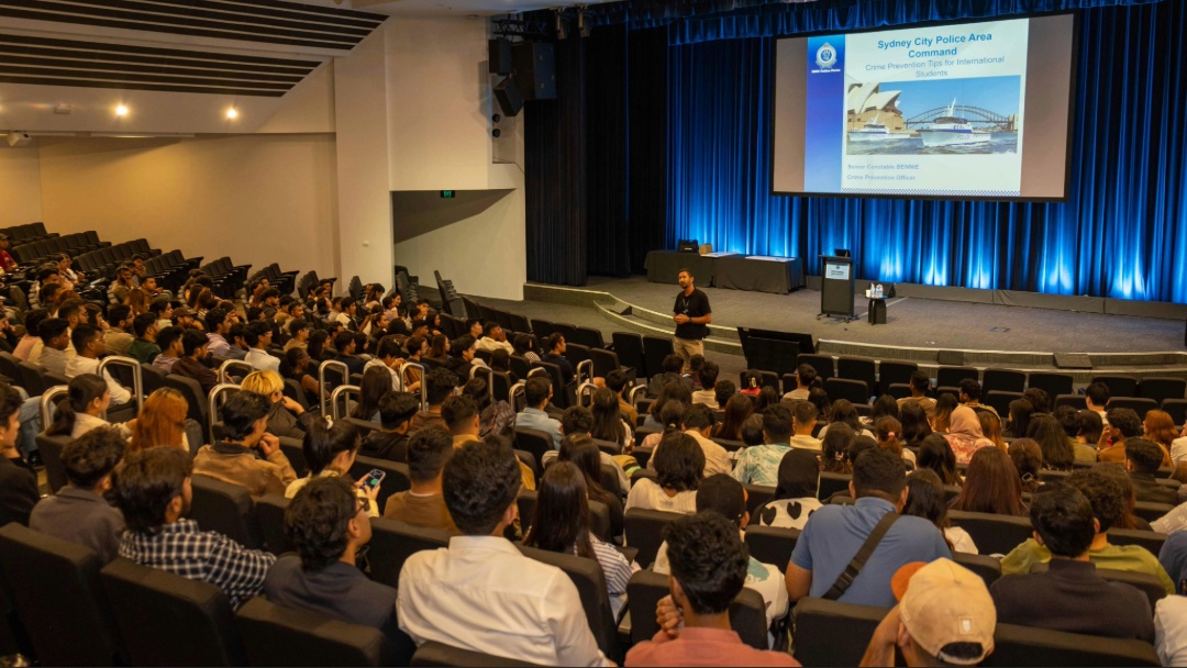 A presenter addresses a full lecture theatre during a crime prevention talk with a presentation slide on screen.