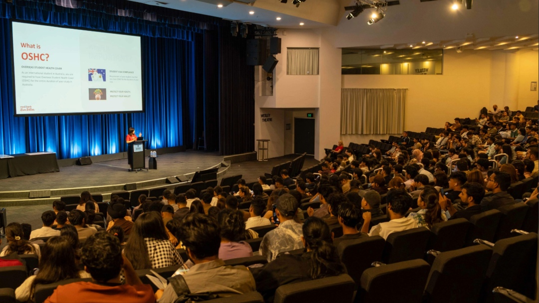 A speaker presents an OSHC health and safety slide to students seated in a large theatre.