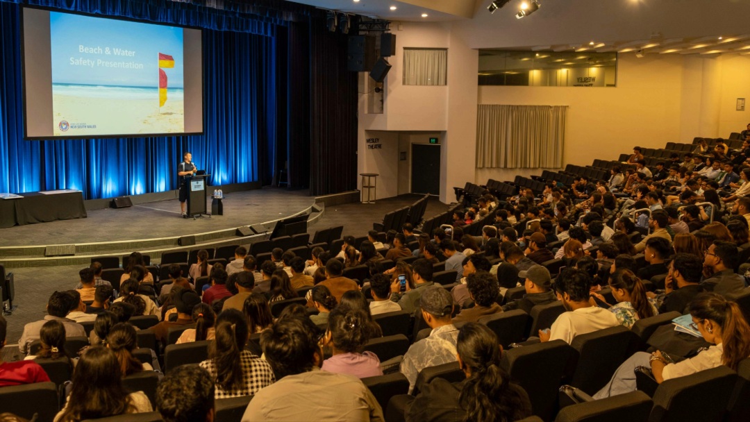 A presenter speaks on stage about beach and water safety to a large audience in a theatre.
