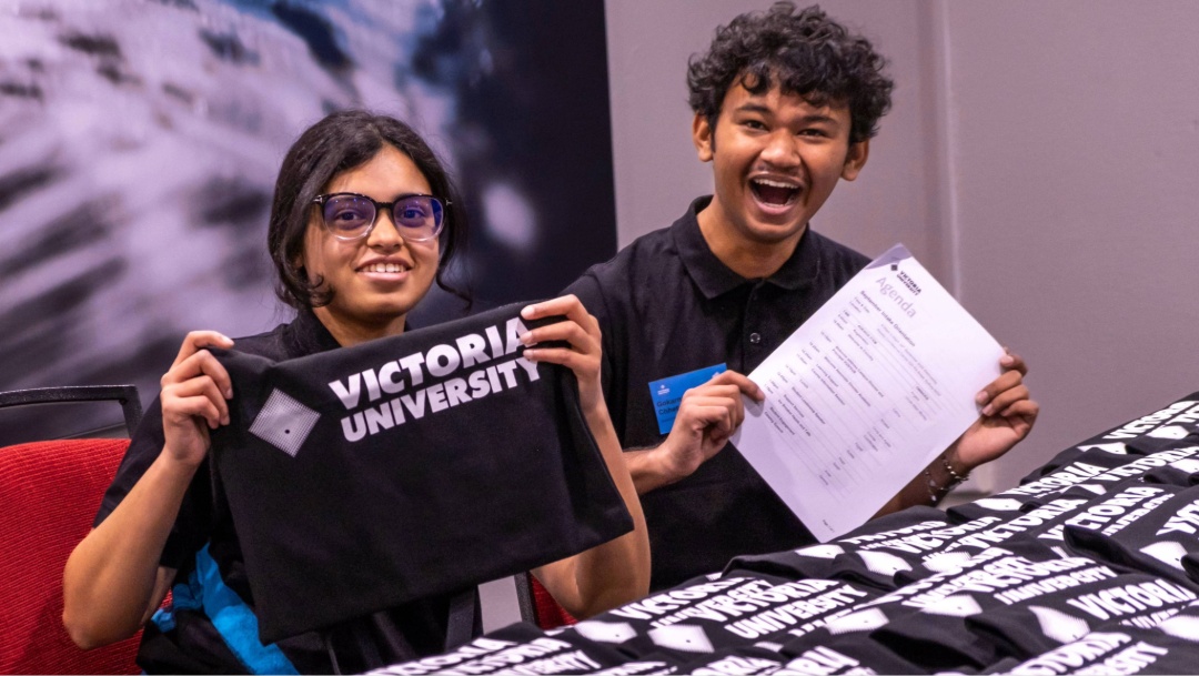 Two students stand behind a table holding Victoria University welcome bags and orientation materials.