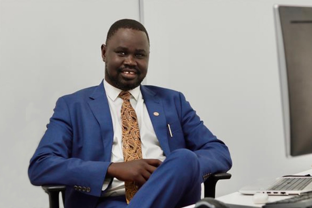 A man wearing a navy suit sitting down in a chair in an office, looking at the camera