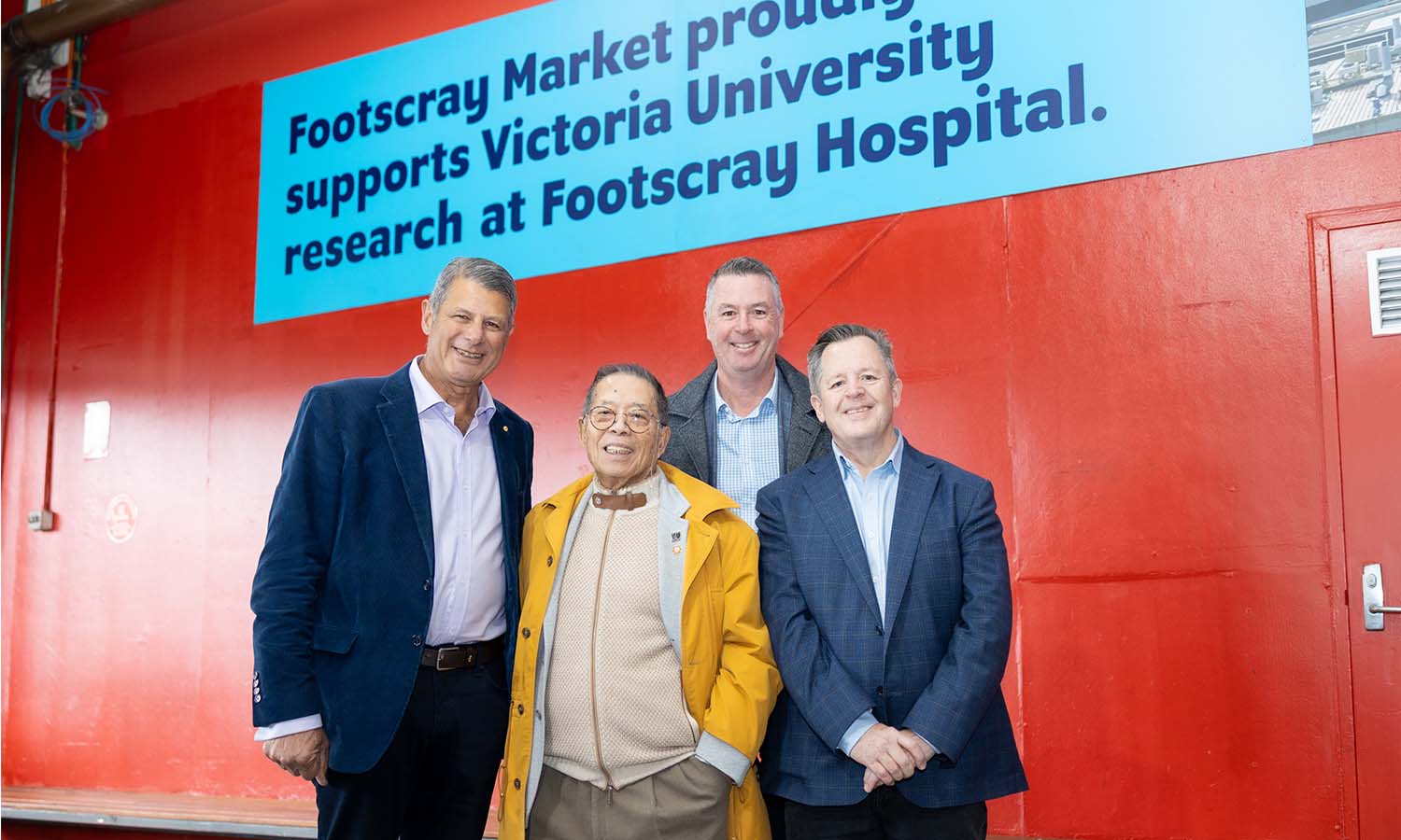 Four men in suits standing in front of a red wall at Footscray Market