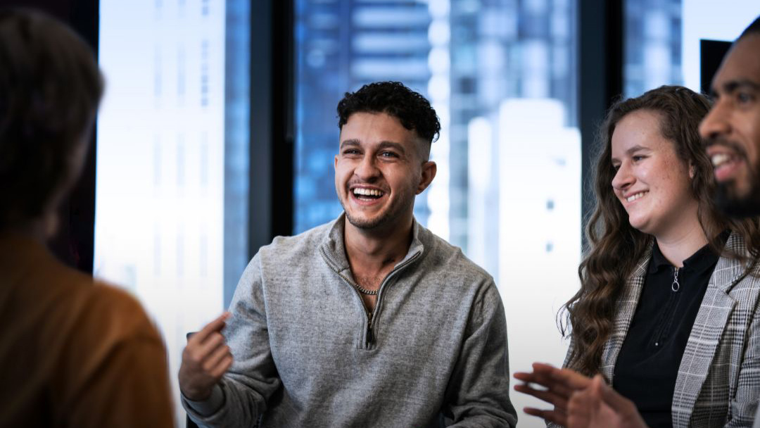 A small group of people standing indoors during a discussion. One person in the foreground is smiling and gestures while speaking, with other participants nearby. Warm indoor lighting and blurred city buildings are visible in the background.