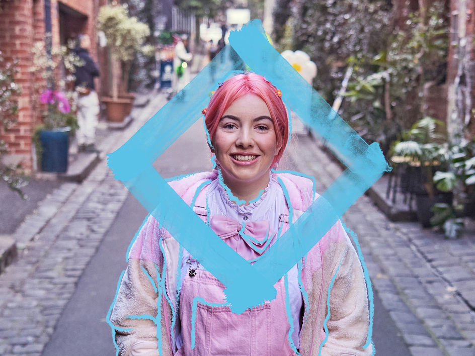 Person standing in the centre of a laneway, wearing pastel pink overalls, a light-coloured jacket. A large blue diamond-shaped frame is positioned around the person's upper body, creating a graphic focal point. The laneway is lined with brick buildings, potted plants and greenery, with the paved street extending into the background. 