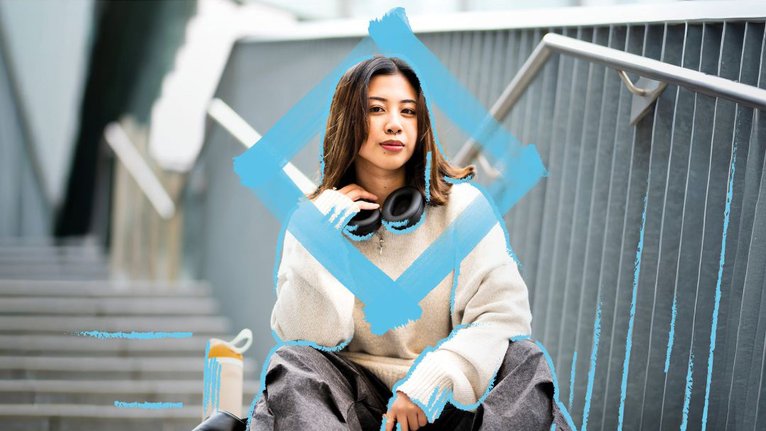 Person seated on outdoor concrete steps wearing large headphones, a white jumper and casual trousers. A large blue diamond-shaped frame is positioned around the person's upper body. A backpack rests nearby, with railings and landscaped campus surroundings visible in the background.