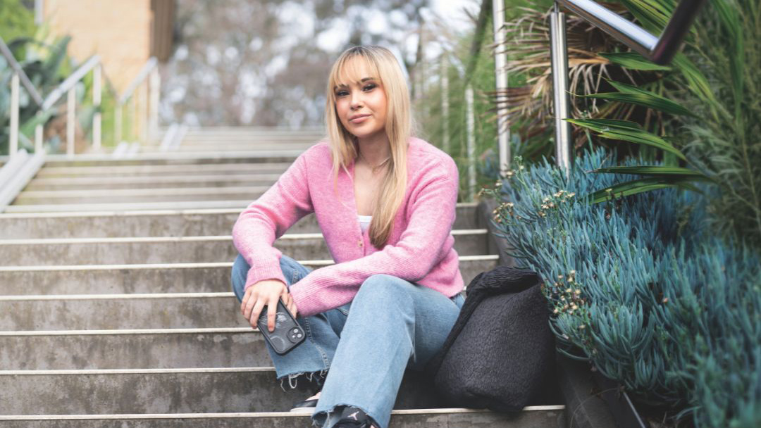 Person seated on outdoor concrete steps wearing a pink top and dark pants, with a backpack beside them. Trees and landscaped greenery appear in the background of a campus setting.