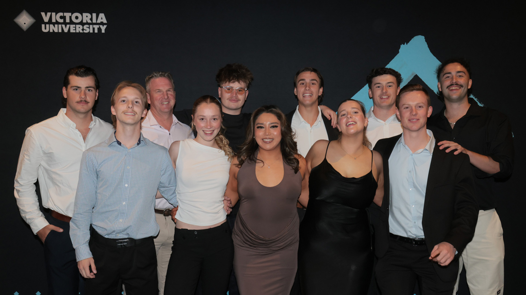 A group of 11 smartly dressed people pose in front of black indoor wall. Victoria University logo is visible in background.