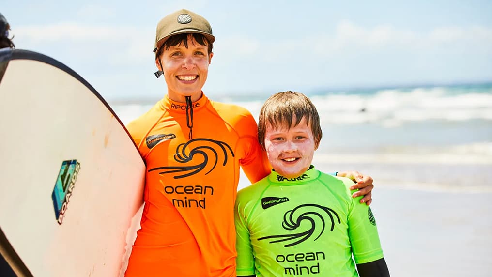 Smiling Ocean Mind surf mentor and young participant standing together on the beach with a surfboard after a surfing session.