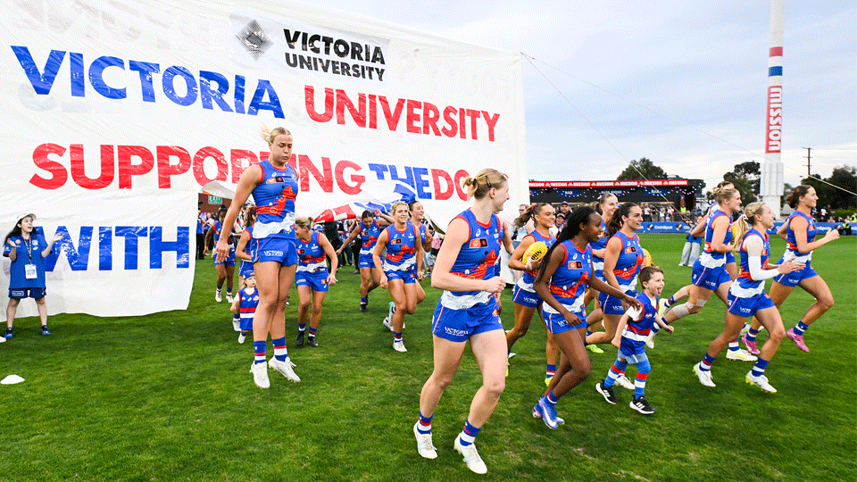Bulldogs AFLW players run onto the ground, through a banner with the words 'Victoria University supporting the Dogs with Pride'