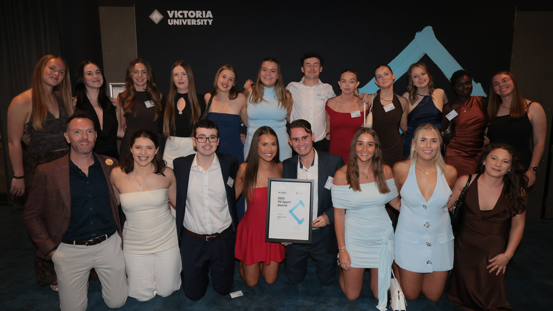 A large group of smartly dressed people pose in front of black indoor wall. Victoria University logo is visible in background.