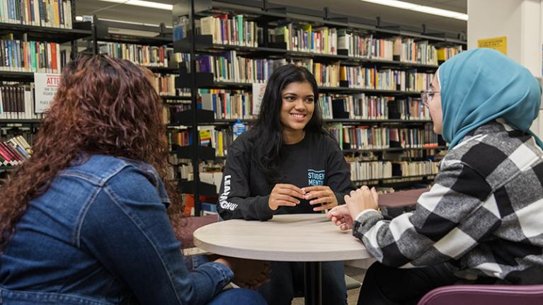study group of 3 multicultural female students in library talks around a table