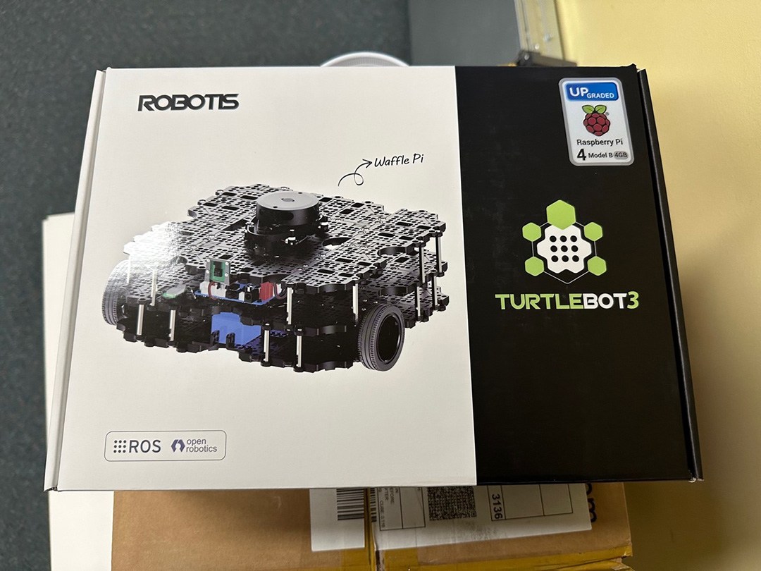 A closed cardboard box. The box has a picture of a small square robot with wheels. The box says "Robotis" and "Turtlebot3" with a Raspberry Pi logo.