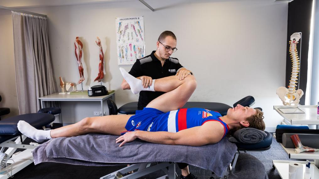 A person in a black & white uniform massages a man wearing a blue and white sport uniform in a room with anatomical posters and models