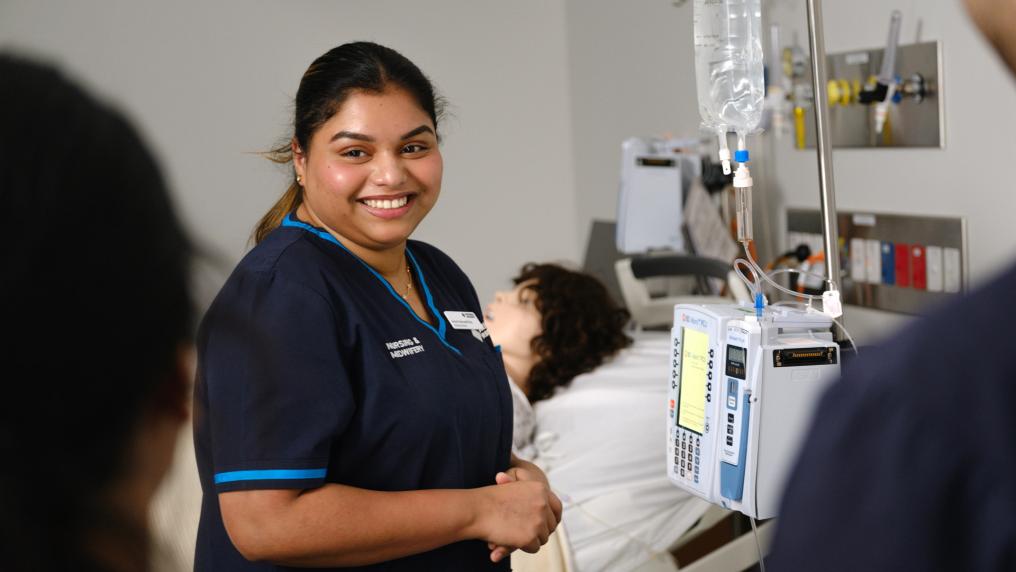 A nursing student standing next to a simulation hospital bed. There's a dummy human with a wig lying in the bed behind the student. The student is smiling is smiling towards two other students standing out of focus in front of the camera.