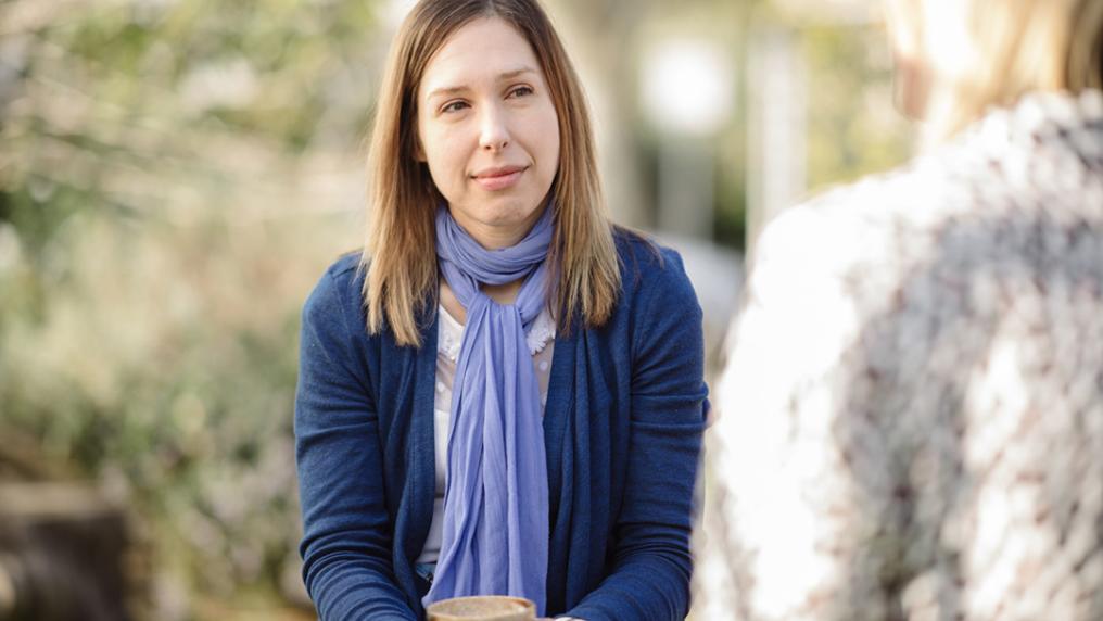 A middle aged woman in a purple scarf looks serious as she speaks with someone