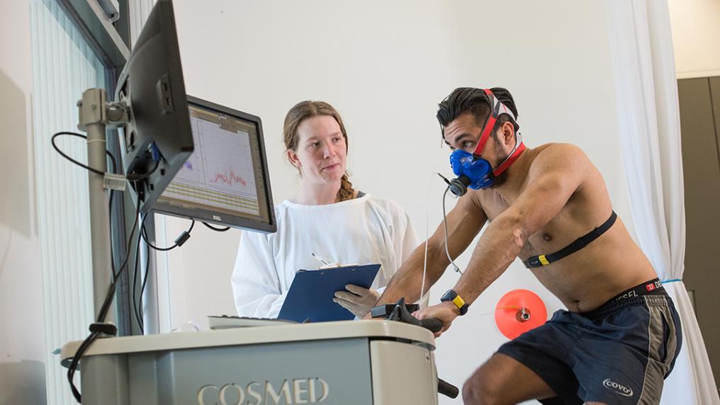 man hooked up to monitoring equipment rides an exercise bike, watched by a woman in a lab coat