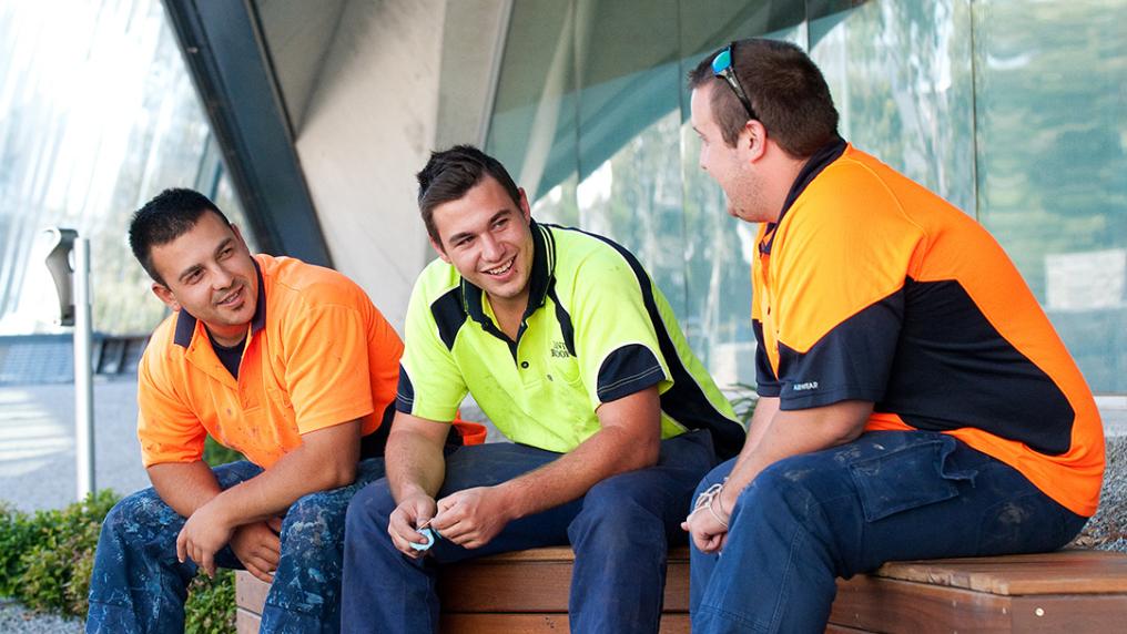 three young men in trades uniforms chat outside a modern campus building