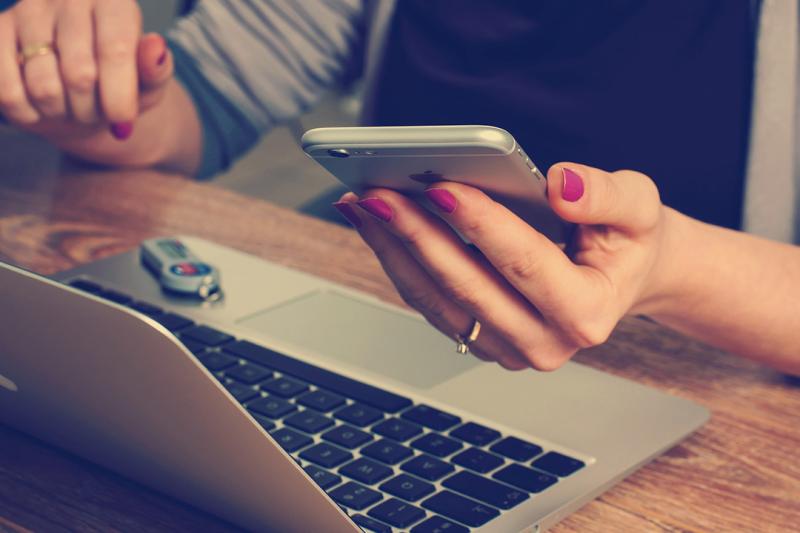 A hand holds a phone while hovering over a laptop