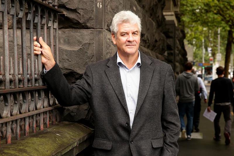 An older man in a relaxed suit poses on a treelined street