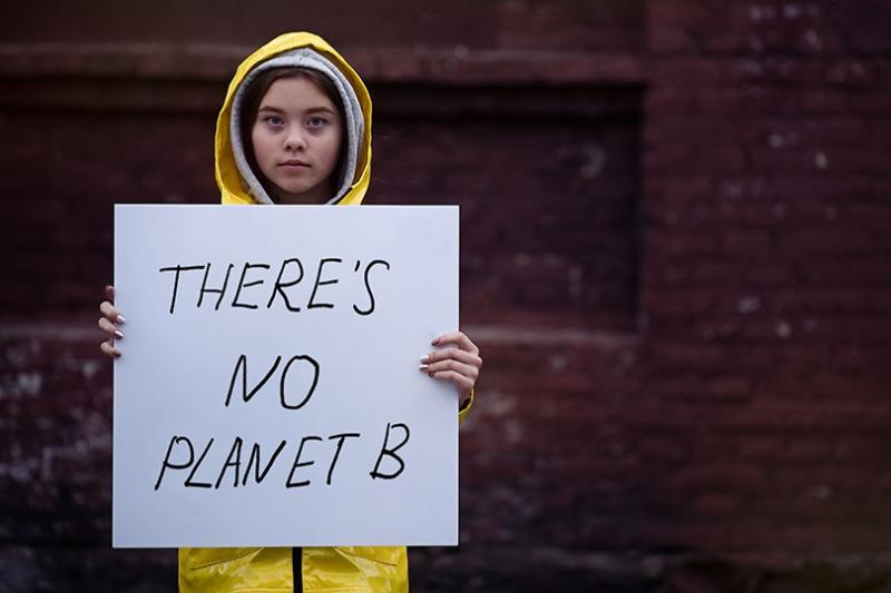 A young girl in a yellow raincoat holds a sign reading 'There's no planet b'