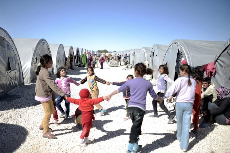 A group of children hold hands in a circle in what looks like a game, in an arid tent community