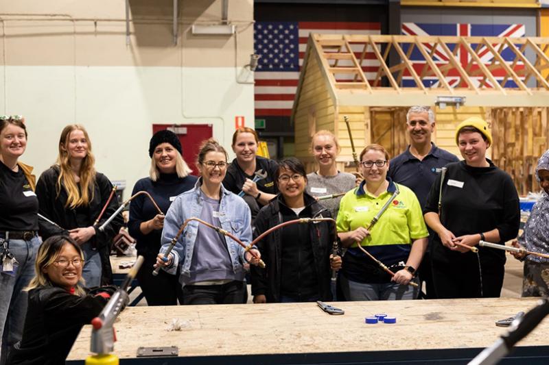 A multicultural group of women holding copper pipe and tools at a work bench, smiling