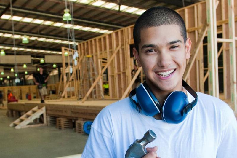 A carpentry student with ear protection around their neck. The student is holding a hammer. In the background is a house frame being built.