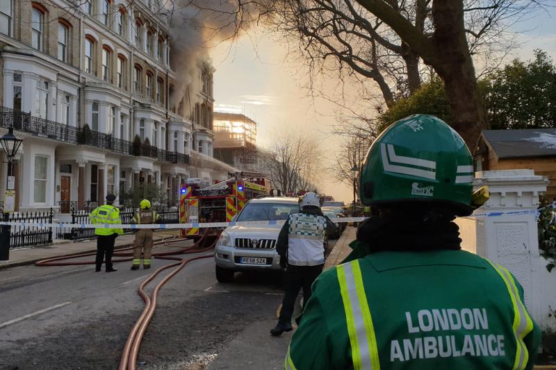 A person wearing a London Ambulance uniform standing outside. In the background are apartment buildings, with one surrounded by smoke.