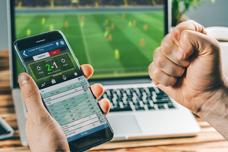 A person holding a phone with a betting app on it, watching a sports game on a laptop.
