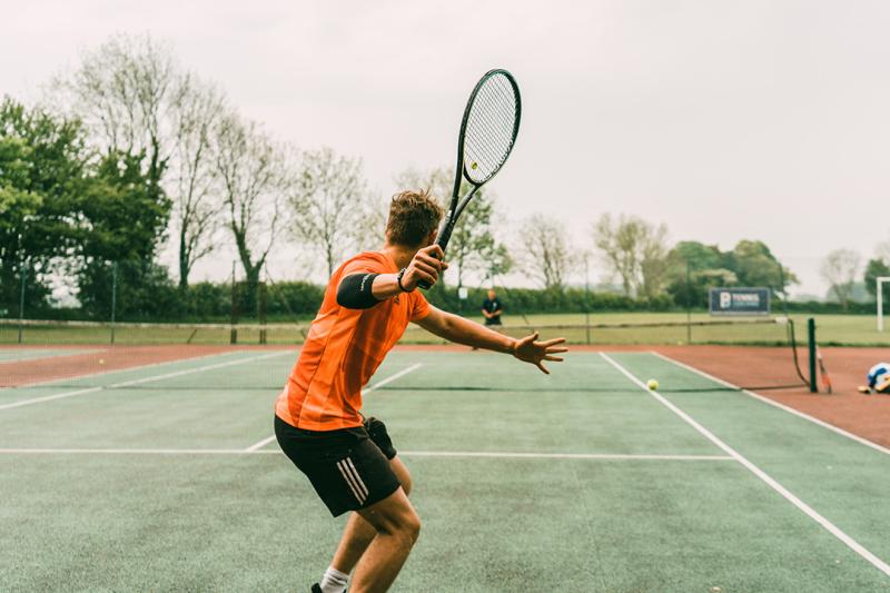 A man in orange sport uniform plays tennis on an asphalt court