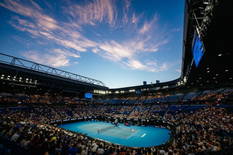 A tennis court arena. The arena is filled by a crowd of spectators. The sky is dark blue, as the sun is setting. The court is illuminated by artificial lights.