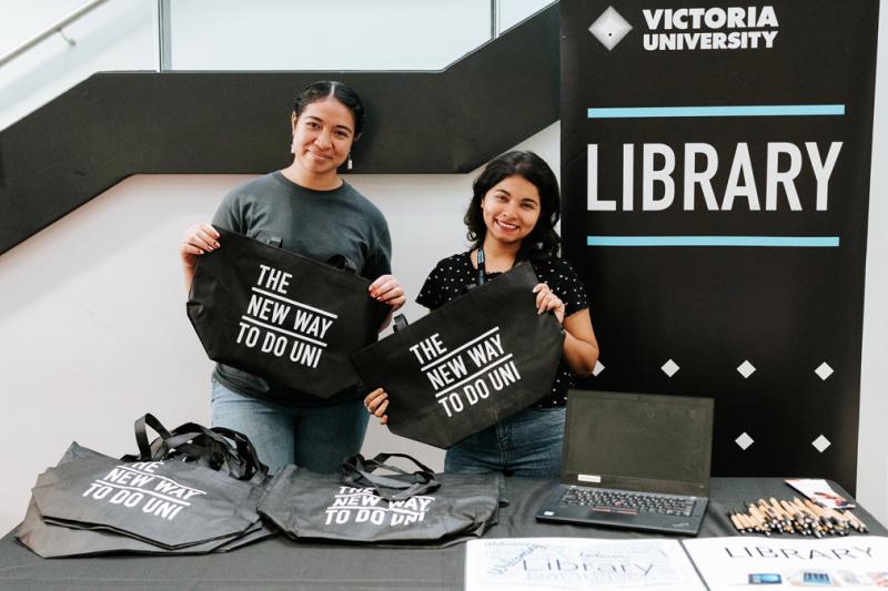 Two students in Victoria University shirts standing behind a table at a stall. The stall has various papers and bags.