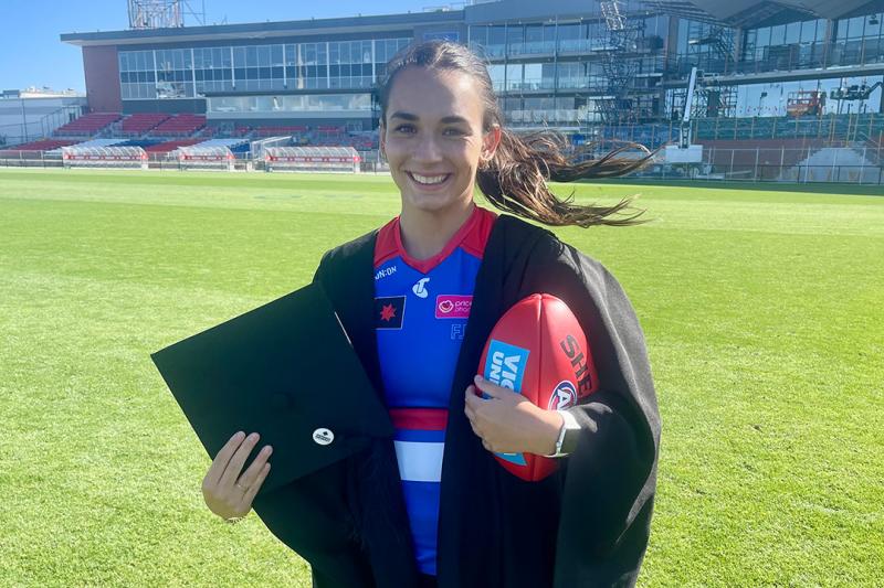 A young woman in a bule, white and red sport uniform and graduation gown, holds a football