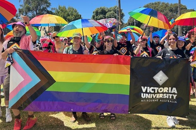 A group of more than 40 people march behind a Pride Flag, in gardens in St Kilda. It is a sunny day and the marchers look happy, wearing rainbow colours and carrying rainbow parasols.