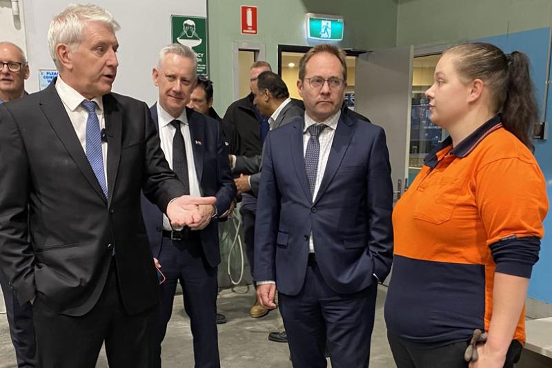 A group of white men in suits talk to a young woman in a trades uniform