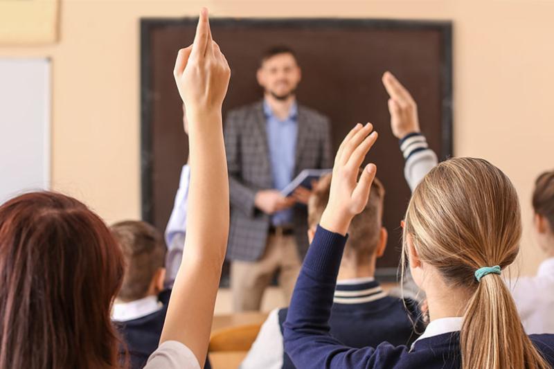 A secondary teacher stands at the front of a classroom, with students in uniforms raising hands to answer questions.