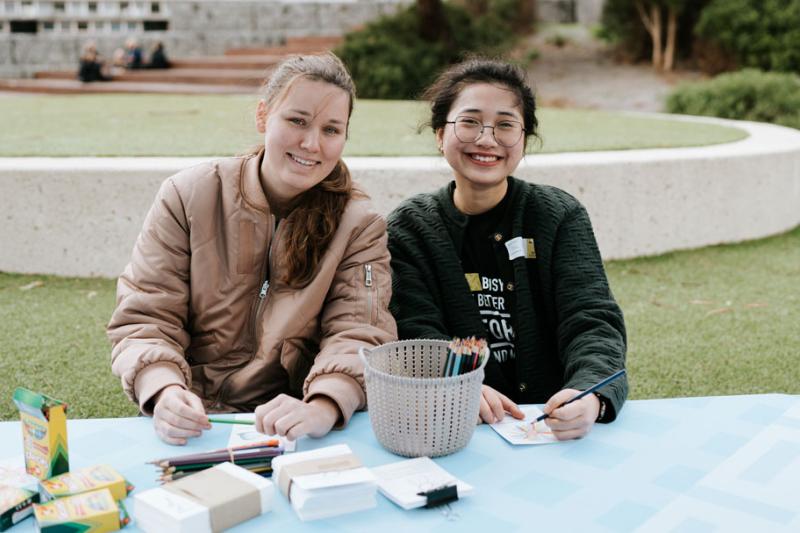 Two students sitting behind a table outside. On the table are some art supplies, including crayons, pencils and paper.