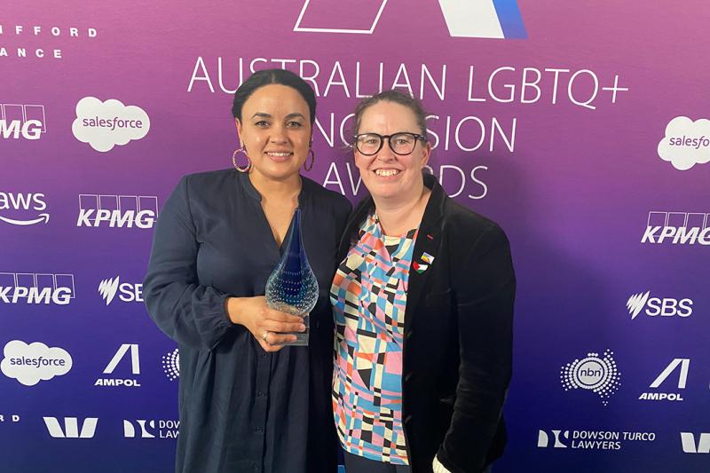 Two women smile and hold a glass trophy, in front of a pink and purple backdrop with sponsorship logos