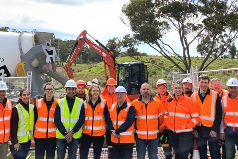 A group of 16 people in hi vis vests smile at the camera, with the trench trial site in background of the photo