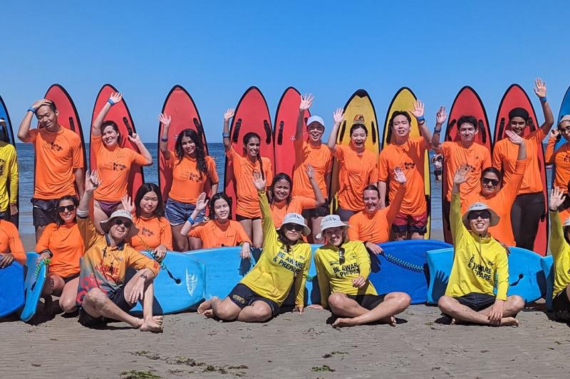 VU students in brightly-coloured swimgear sitting on the beach and waving at the camera.