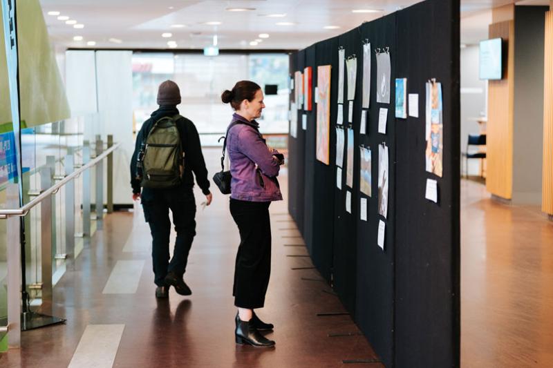 A woman looking at art displays on a temporary wall. The wall is set in the library corridor, with people walking in the background.