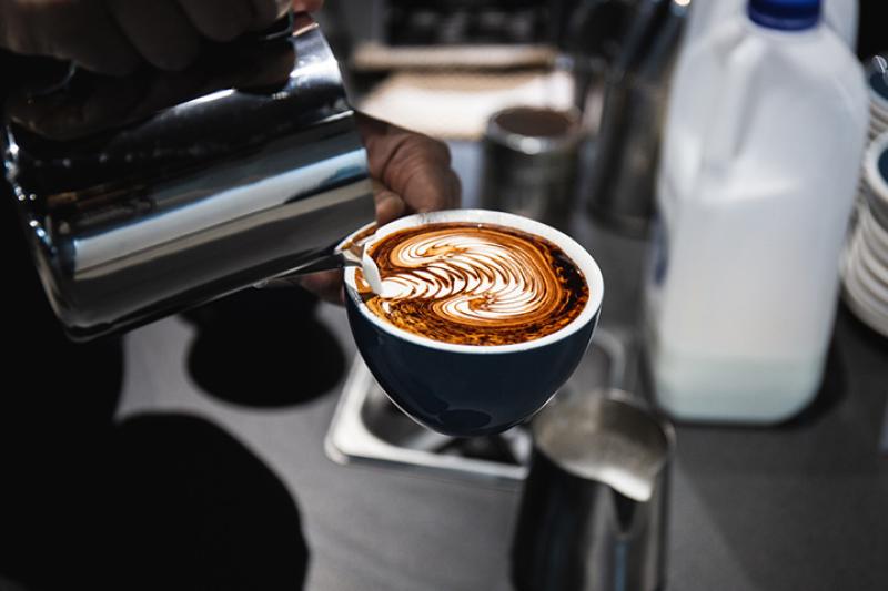 A milk jug is used to create a detailed pattern in milk on a coffee