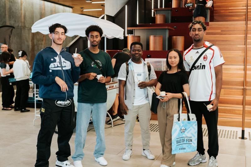 A group of 5 students inside City Campus, looking at the camera. Most of them are holding small cups of ice cream. Behind them is a gelato stall with a white umbrella above it.