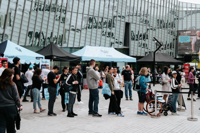 A crowd of people outside P Block at Footscray Park Campus. Around them are a series of outdoor stalls and flags from different companies.