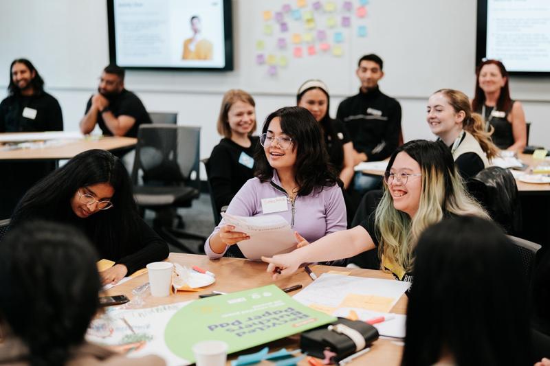 A group of young people in semi-formal clothes laugh around tables while attending a workshop