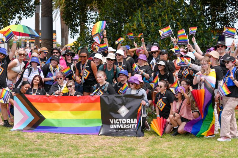 A group of people standing or squatting outside behind a pride flag with the VU logo.