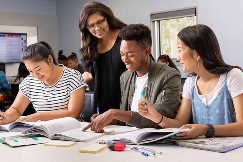 Three VU students sit working in a classroom, while being assisted by a teacher.