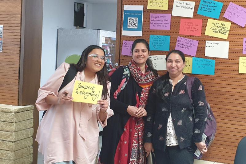 Three students standing in front of a wood panel wall. The wall contains pieces of paper with messages about respect.